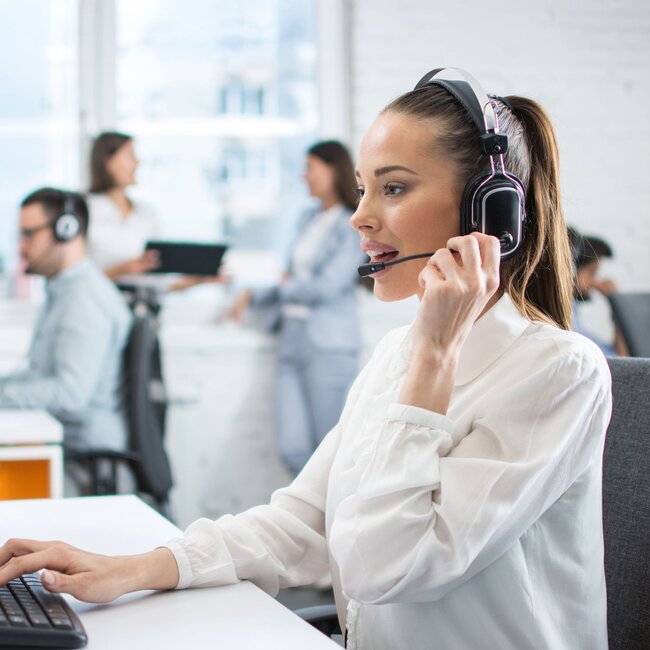 Frau mit Headset im modernen Büro Frau mit Headset arbeitet am Computer im modernen Büro mit Kollegen im Hintergrund