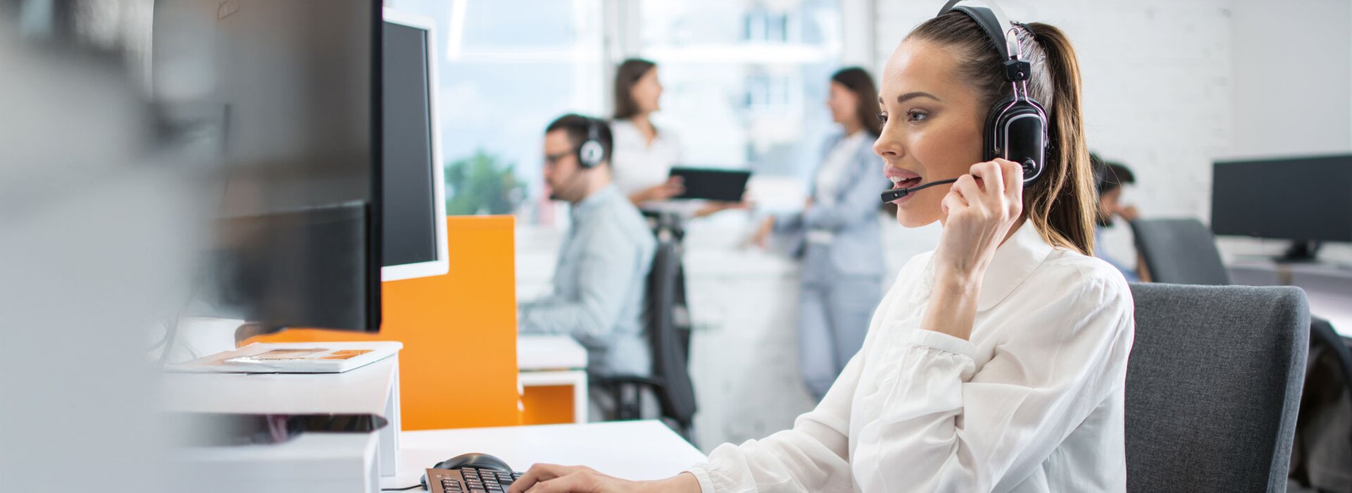 Frau mit Headset arbeitet am Computer im modernen Büro mit Kollegen im Hintergrund