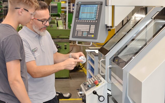 Fachmann erklärt Bedienung einer CNC-Maschine anhand eines Werkstücks
